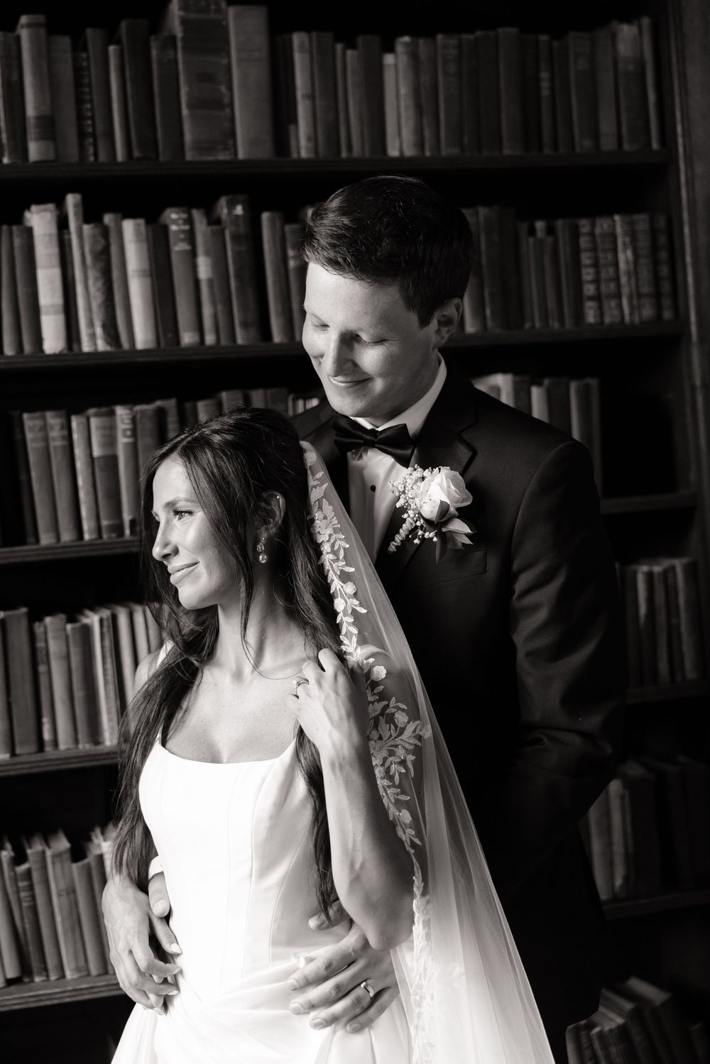 Black and white wedding portrait of bride and groom embracing in front of vintage library bookshelves.