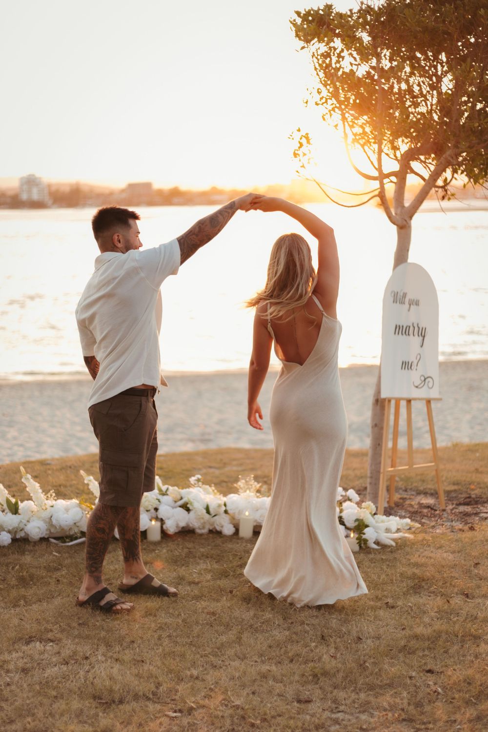 Couple dancing at sunset beach proposal with Will You Marry Me sign and white flowers.