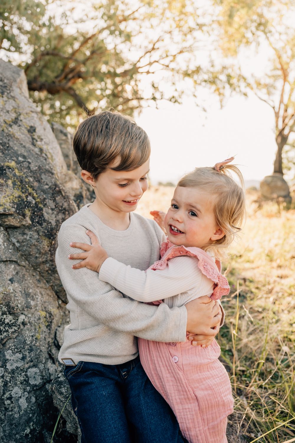 Two children hugging and laughing together while leaning against large rock outdoors.