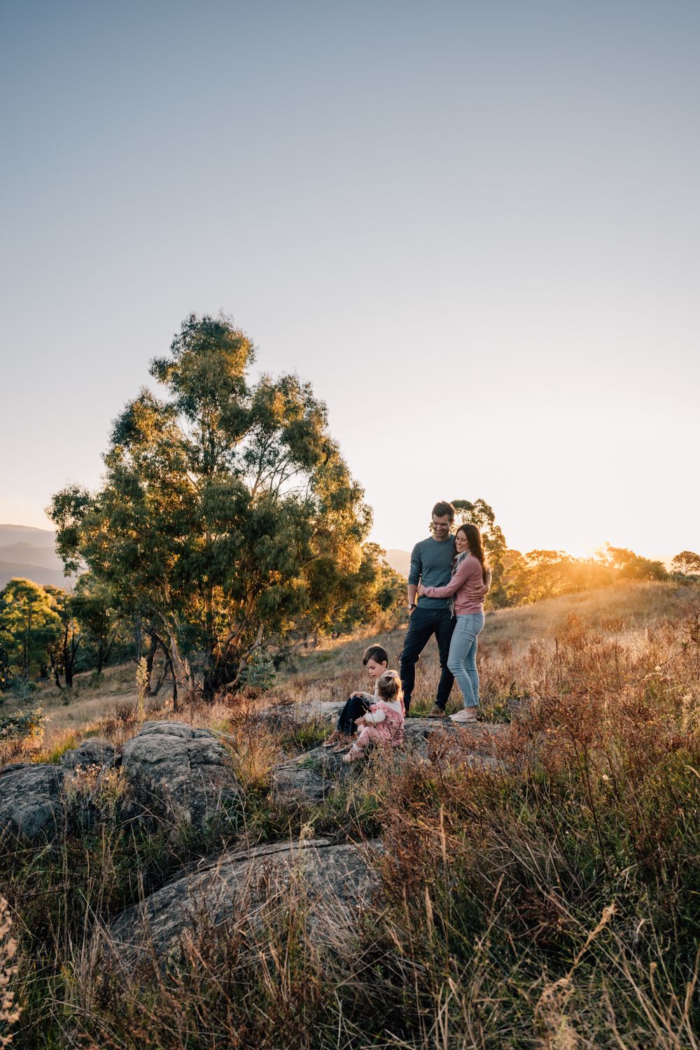 Couple standing on rocky hillside during sunset with dog beside them.