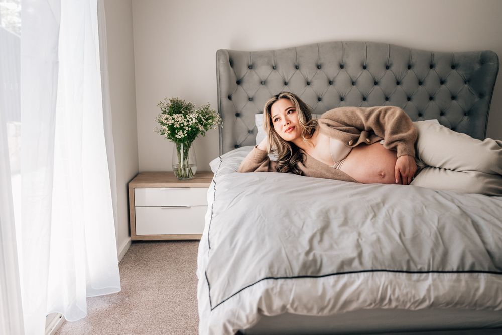 Pregnant woman relaxing on gray tufted bed with white bedding near window.