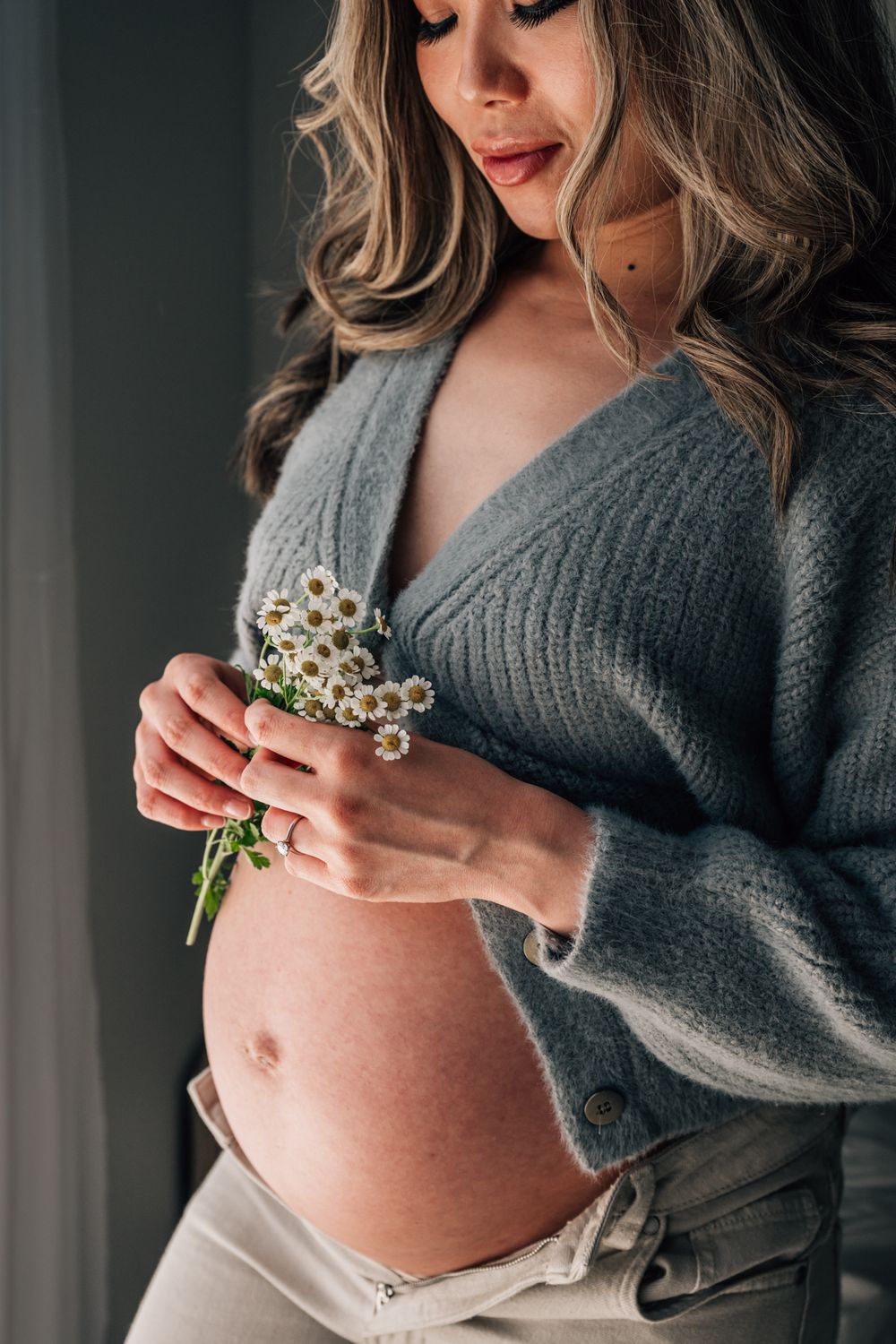 Close up of pregnant belly with woman holding small white flowers in gray sweater.