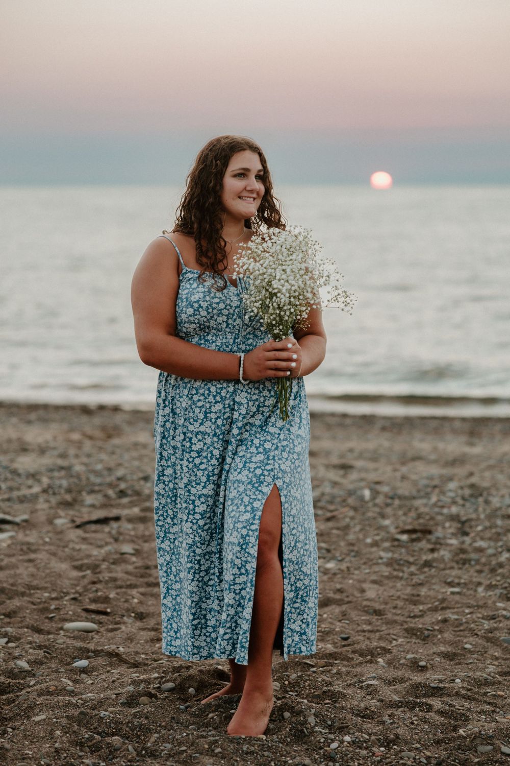 A person in a blue floral dress holds baby's breath flowers on a beach at sunset.