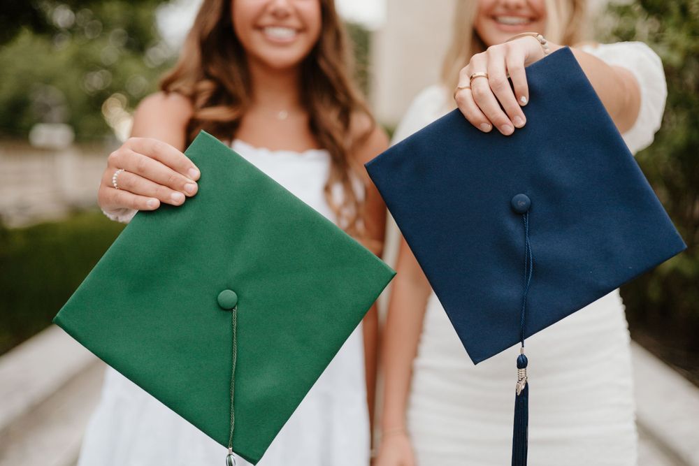 Two graduates holding up their green and navy graduation caps with tassels in an outdoor setting.