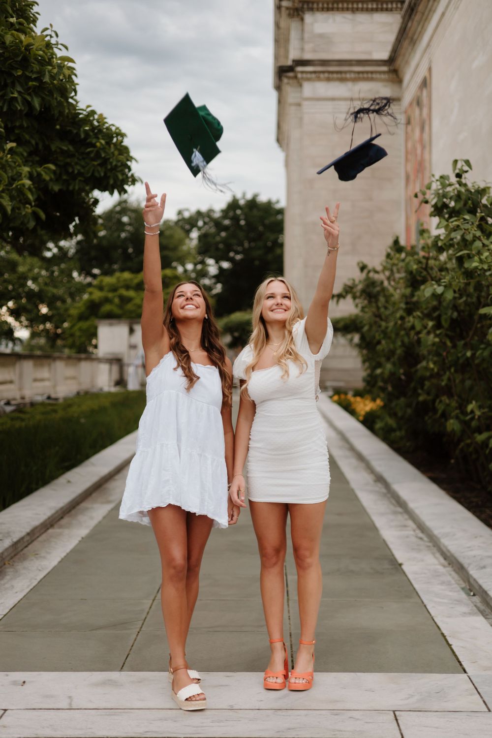 Two friends in white dresses tossing graduation caps in celebration outside a building.