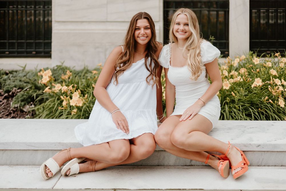 Friends in white dresses pose together on stone steps surrounded by yellow flowers.