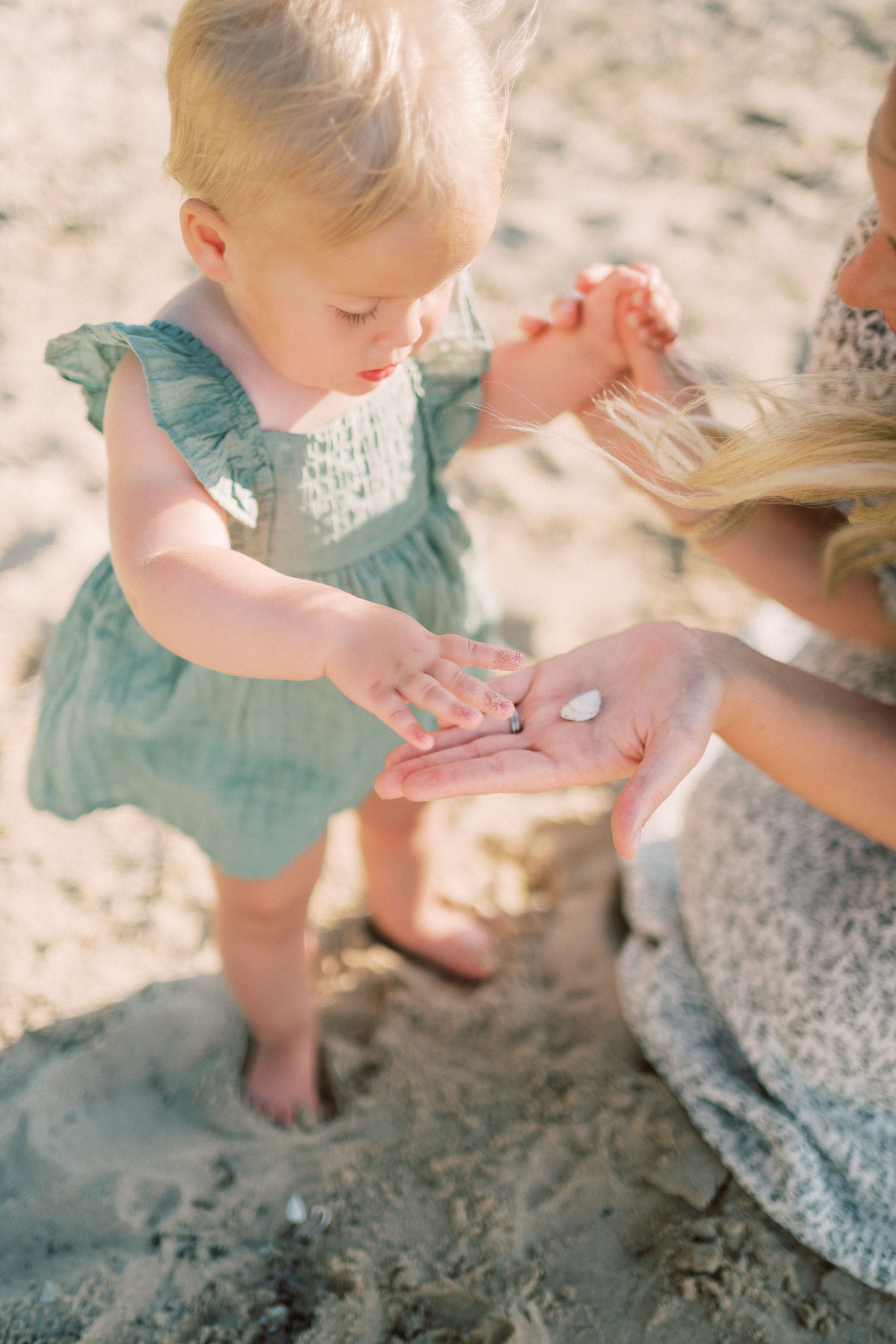 A toddler wearing a teal dress explores the sand on a beach with their hands on a sunny day.