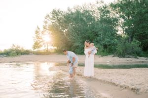 Three people stand at the waters edge on a sandy beach during golden hour sunset with trees in the background.