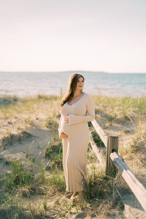 A beige maxi dress flows in the breeze as someone stands on a sandy beach dune overlooking the ocean at sunset.