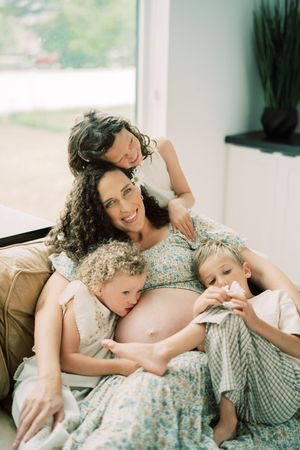 A pregnant woman in a floral dress rests on a couch with two children and another person in a tender family moment.