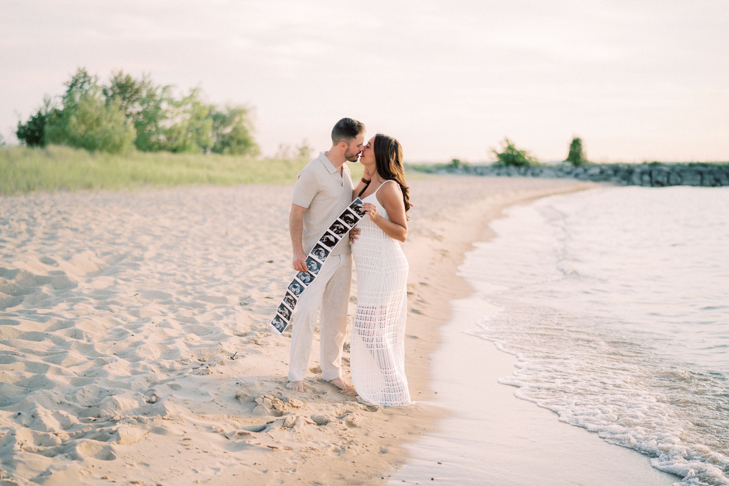 A romantic couple in white clothing embraces on a sandy beach at sunset by the water's edge.