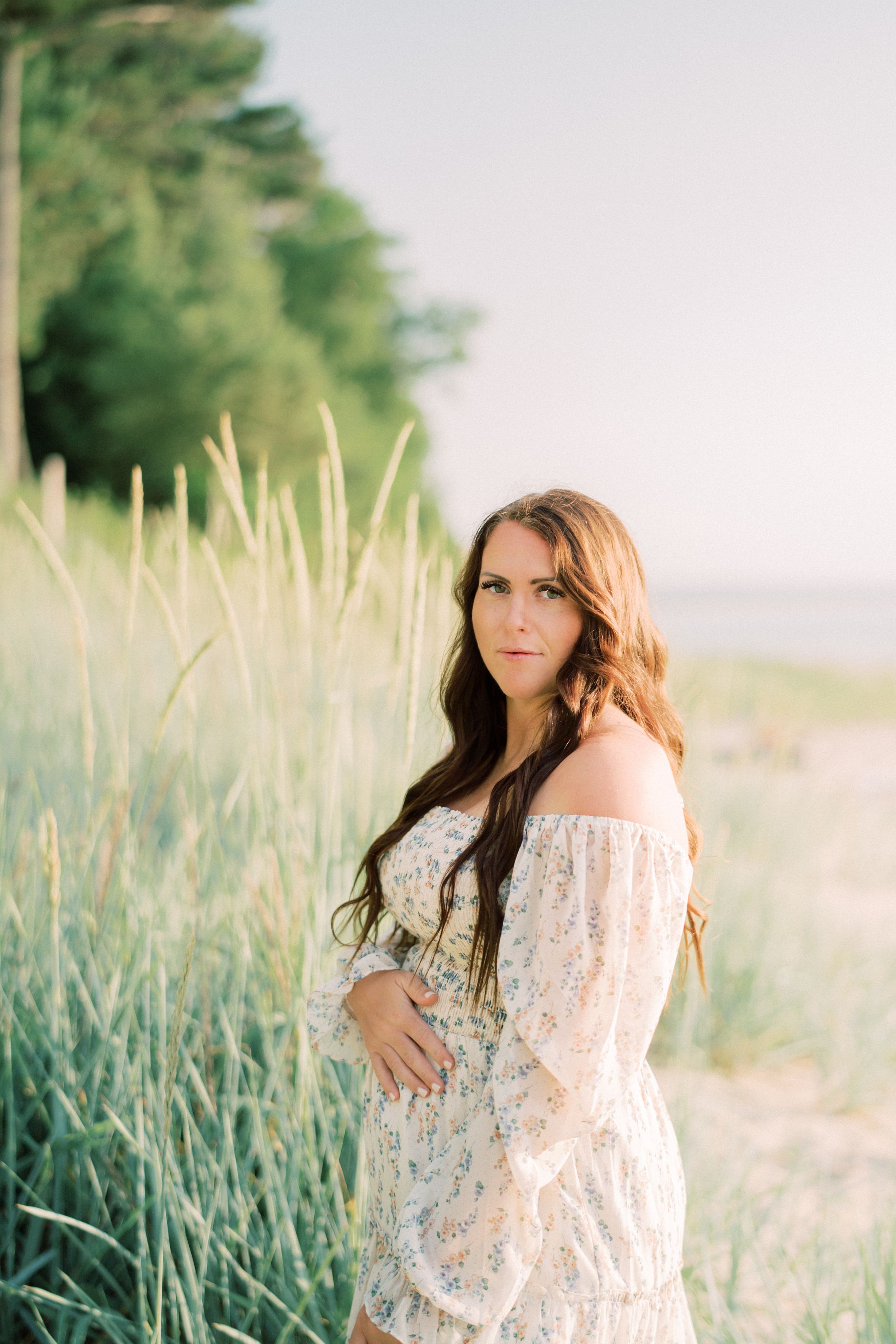 A woman in a delicate white lace dress poses gracefully among tall grass in soft natural light.