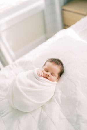 A sleeping newborn baby wrapped in soft white swaddle blanket rests peacefully on white bedding.