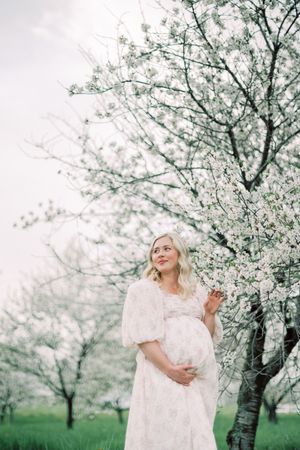 Dreamy maternity portrait shows pregnant woman in white dress standing beneath blooming spring cherry blossom trees.