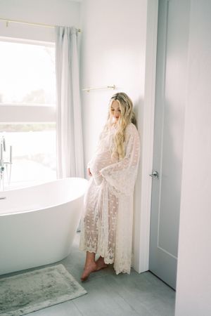 A pregnant woman in white lace robe stands in a bright modern bathroom with freestanding tub and marble floors.