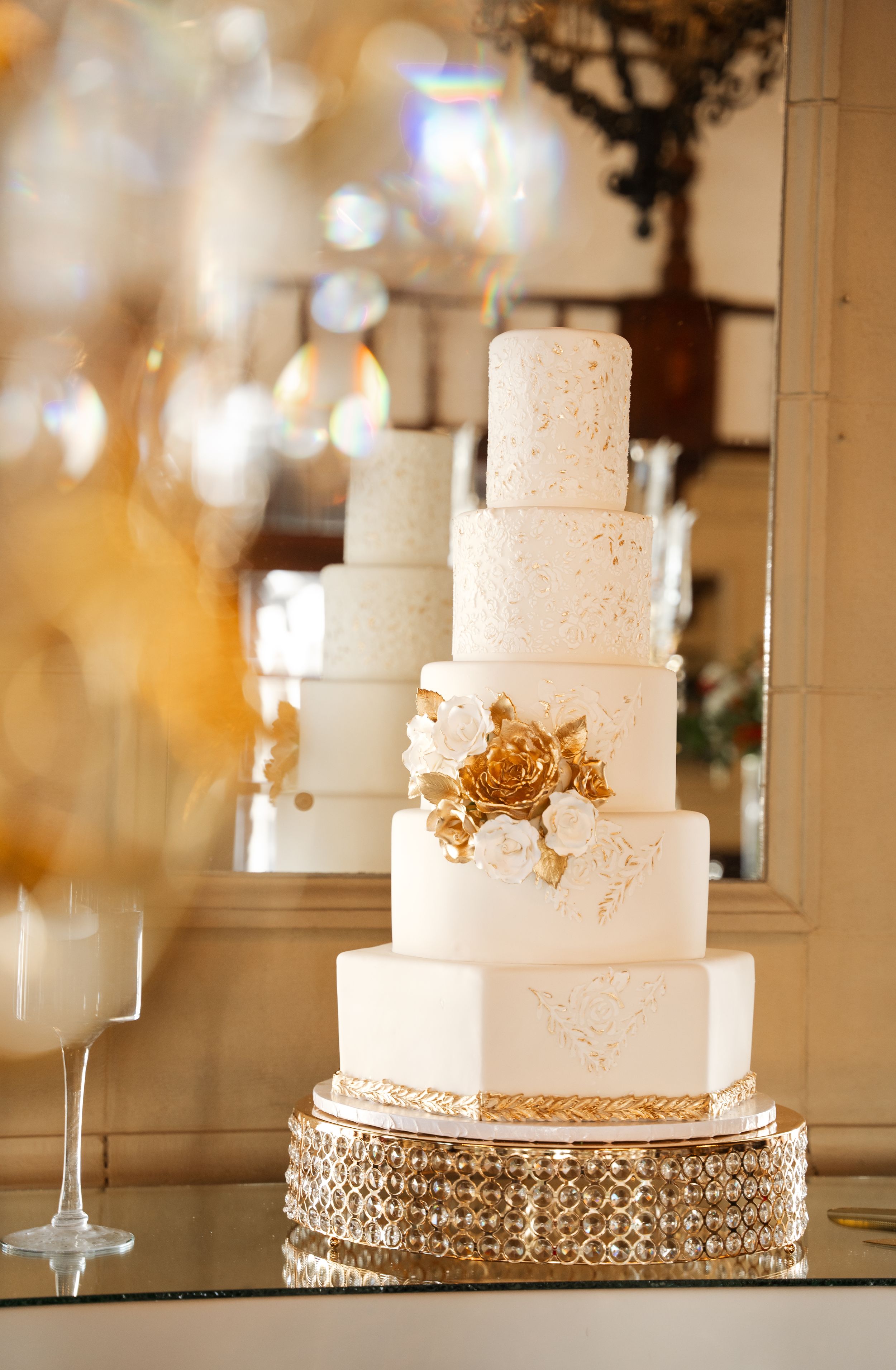 Elegant six-tier white wedding cake with gold accents and floral details on a crystal stand.