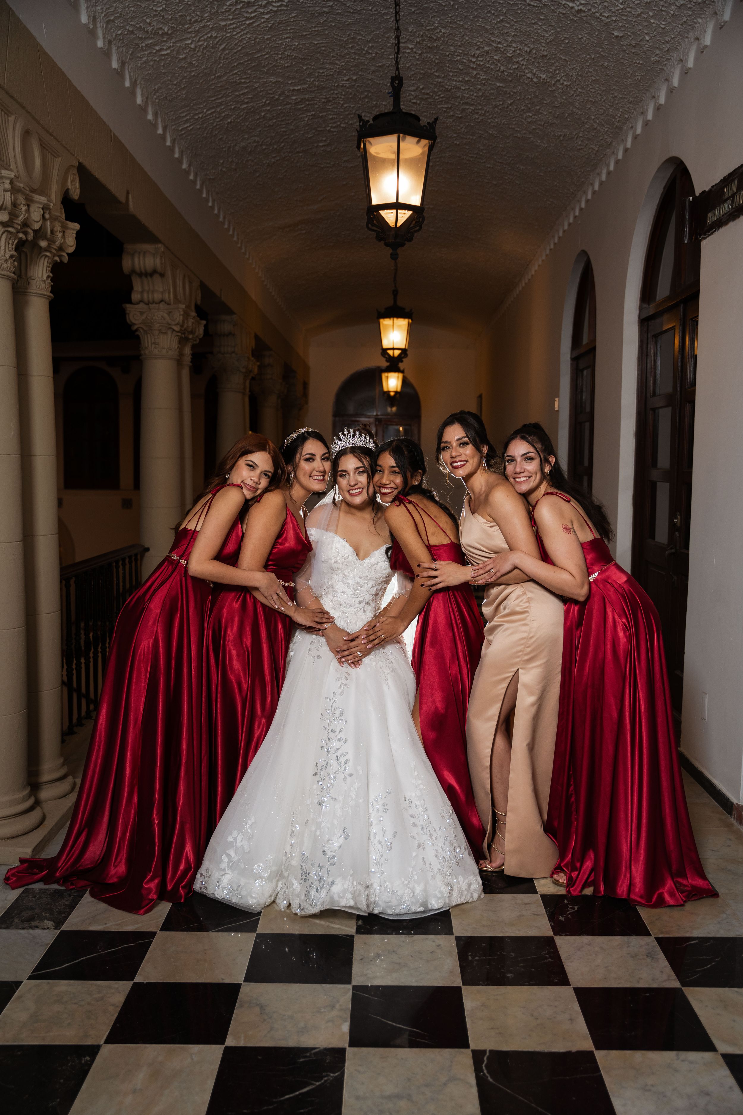 Wedding party poses in elegant hallway with burgundy gowns against checkered floor and archways.
