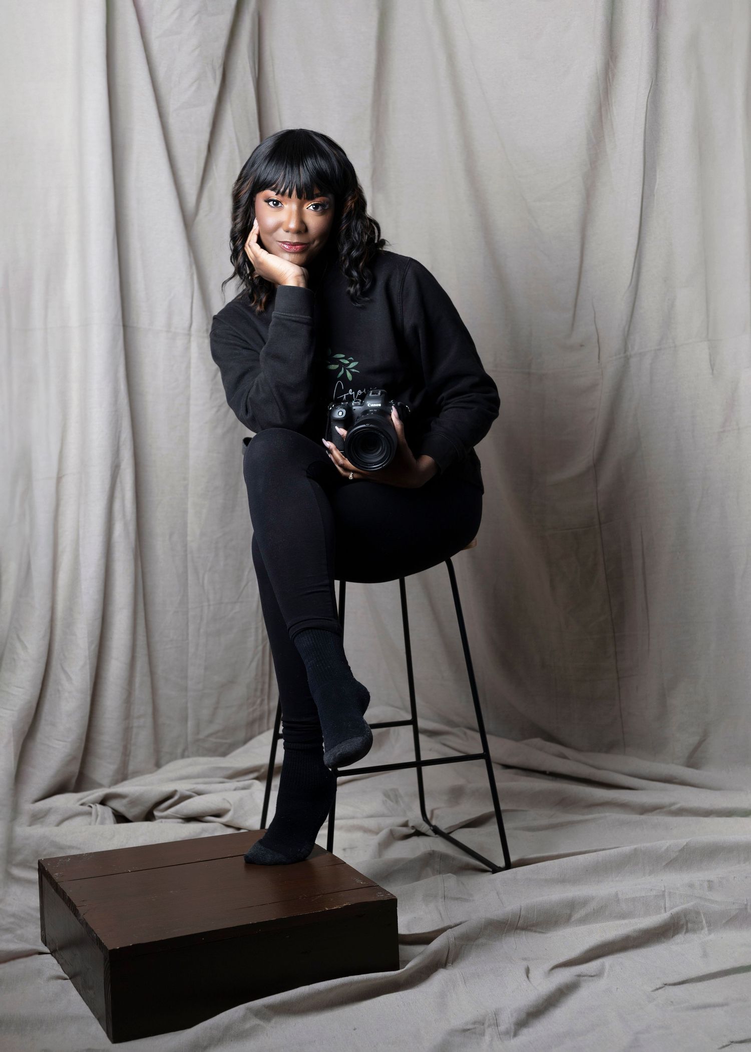 Photographer in black outfit poses with camera against gray studio backdrop.