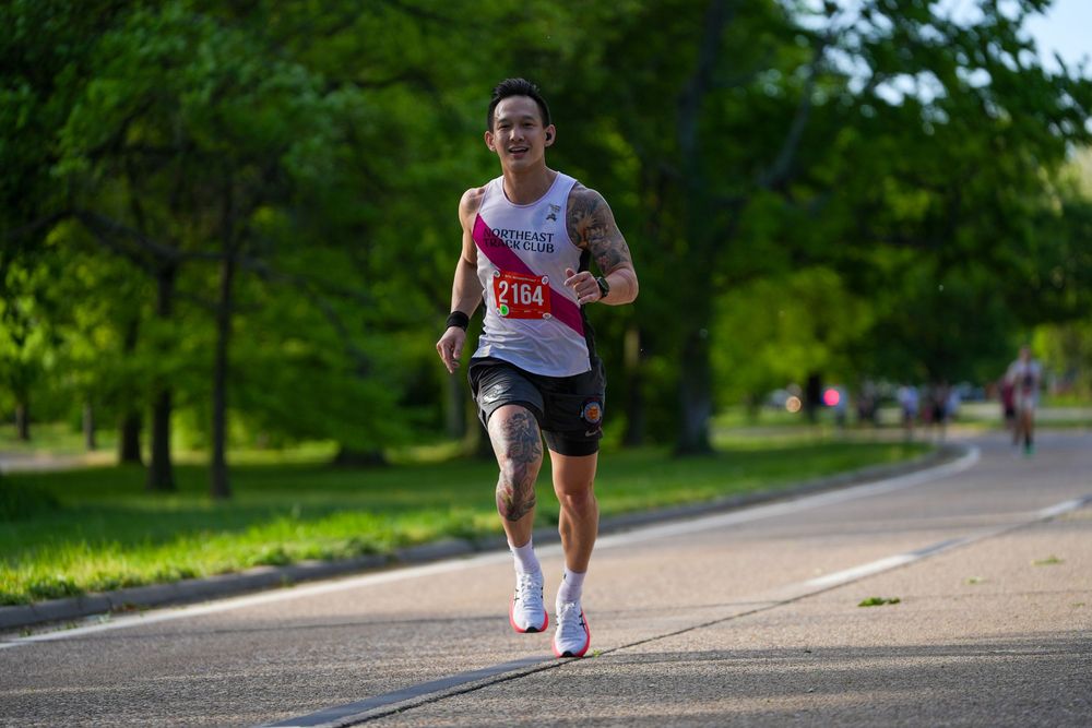 Runner in white tank top and black shorts jogging on path through green park on sunny day.