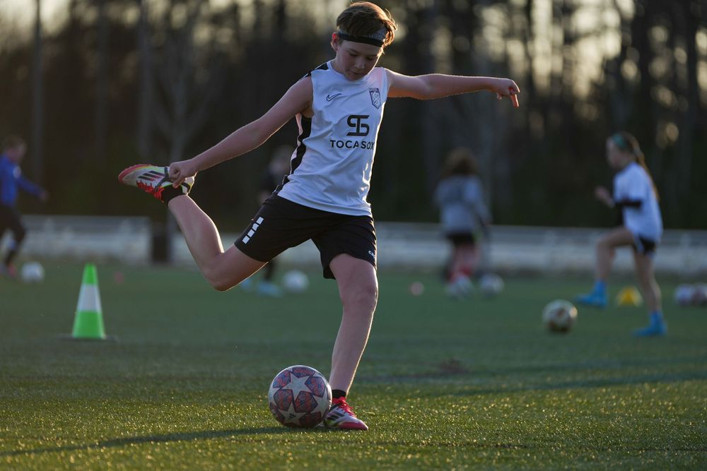 Soccer player in white jersey and black shorts practices kicking technique on grass field during sunset training.