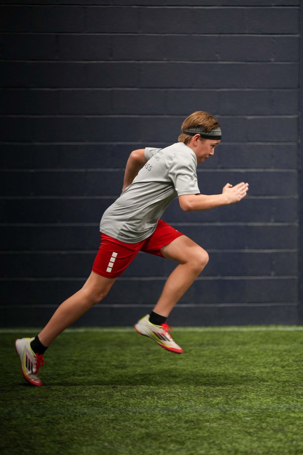 Athlete in gray shirt and red shorts sprinting explosively on indoor turf against dark wall.