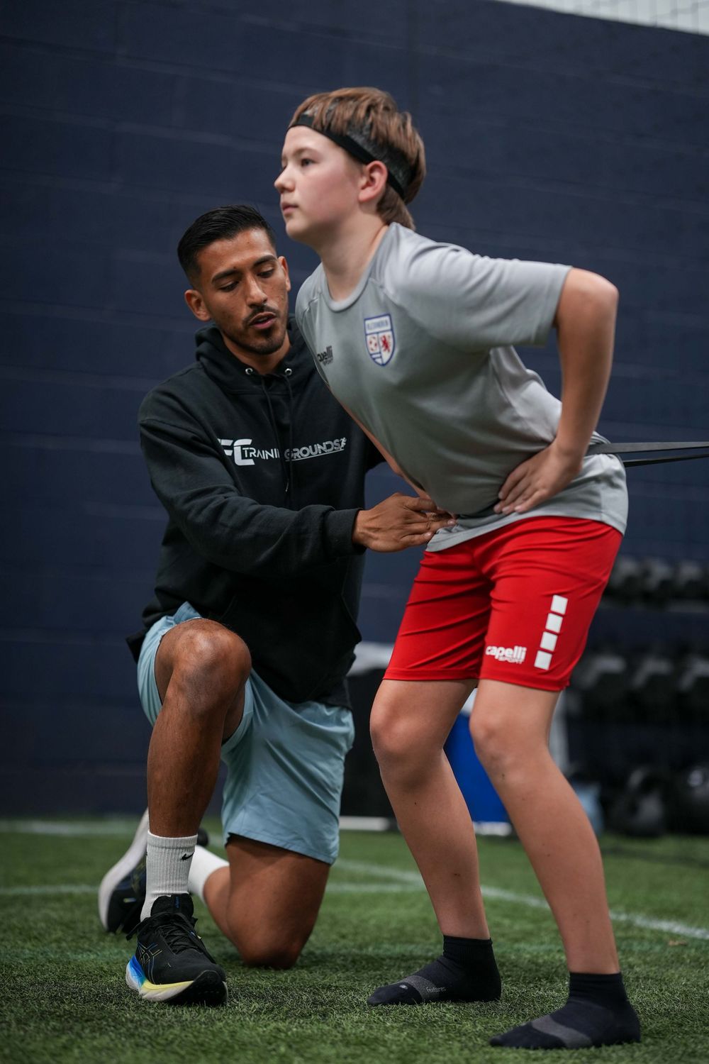 A trainer helps a young athlete with proper form during a coaching session on an indoor turf field.