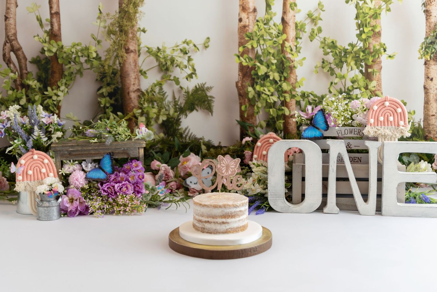 Simple white wedding cake on wooden stand with greenery backdrop and rustic star decorations against light wall.