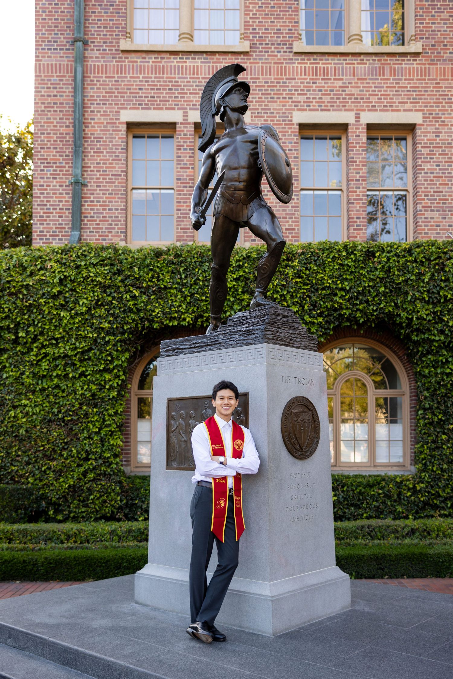 A collegiate statue stands on a stone pedestal in front of a brick building with manicured hedges.
