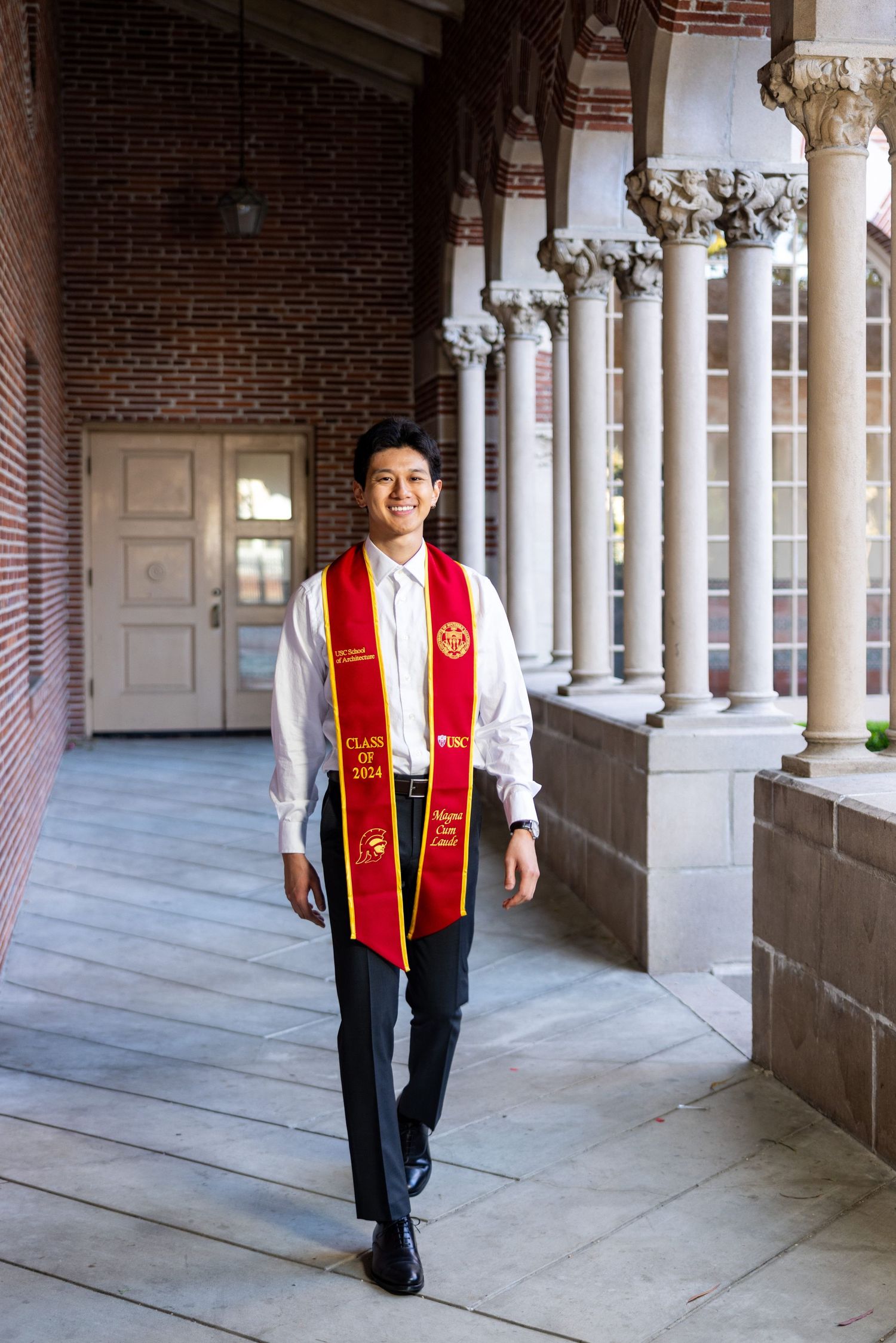 A graduate walks through an ornate columned hallway wearing academic regalia and stoles.