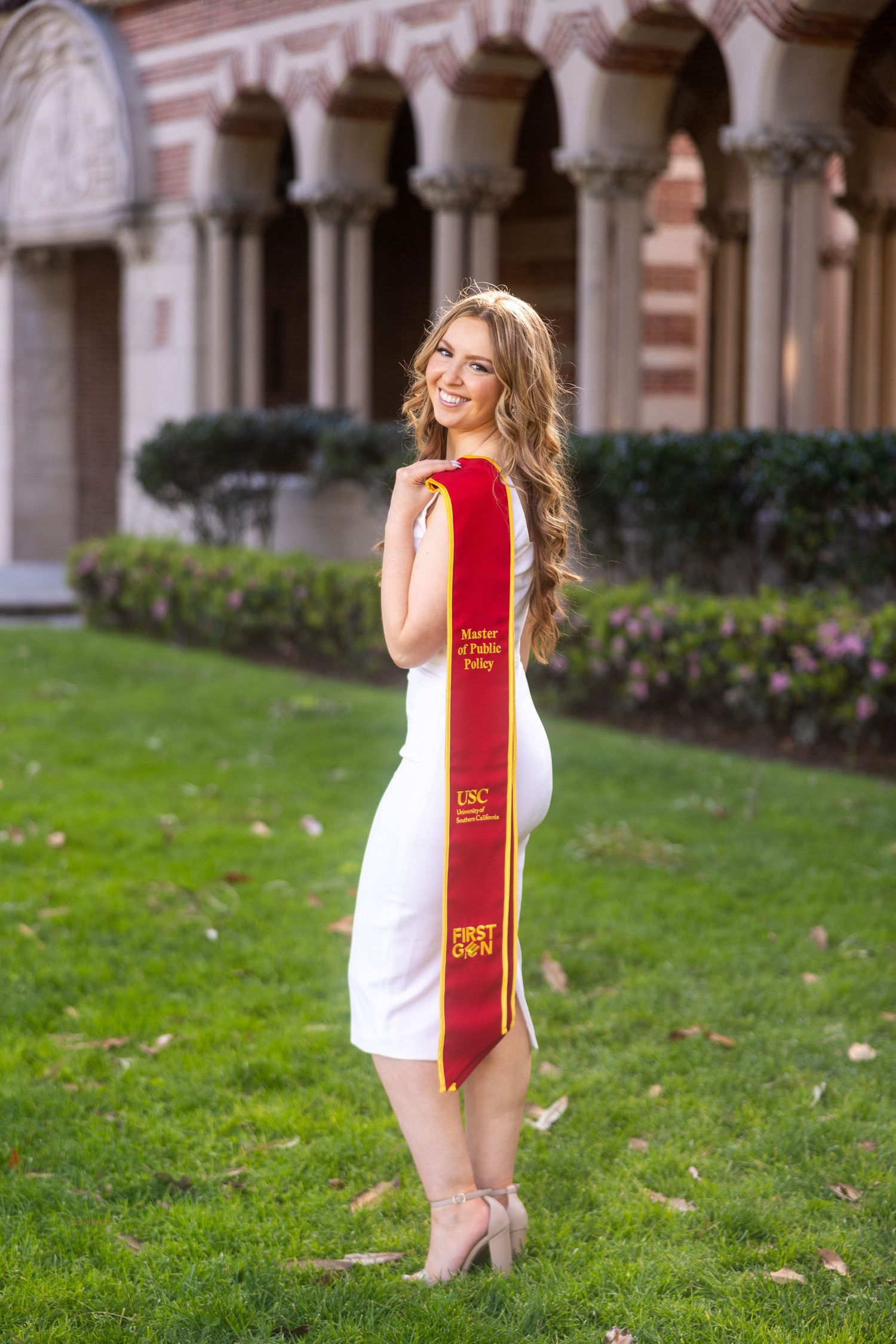 Graduate in white dress and red sash poses on college campus lawn.