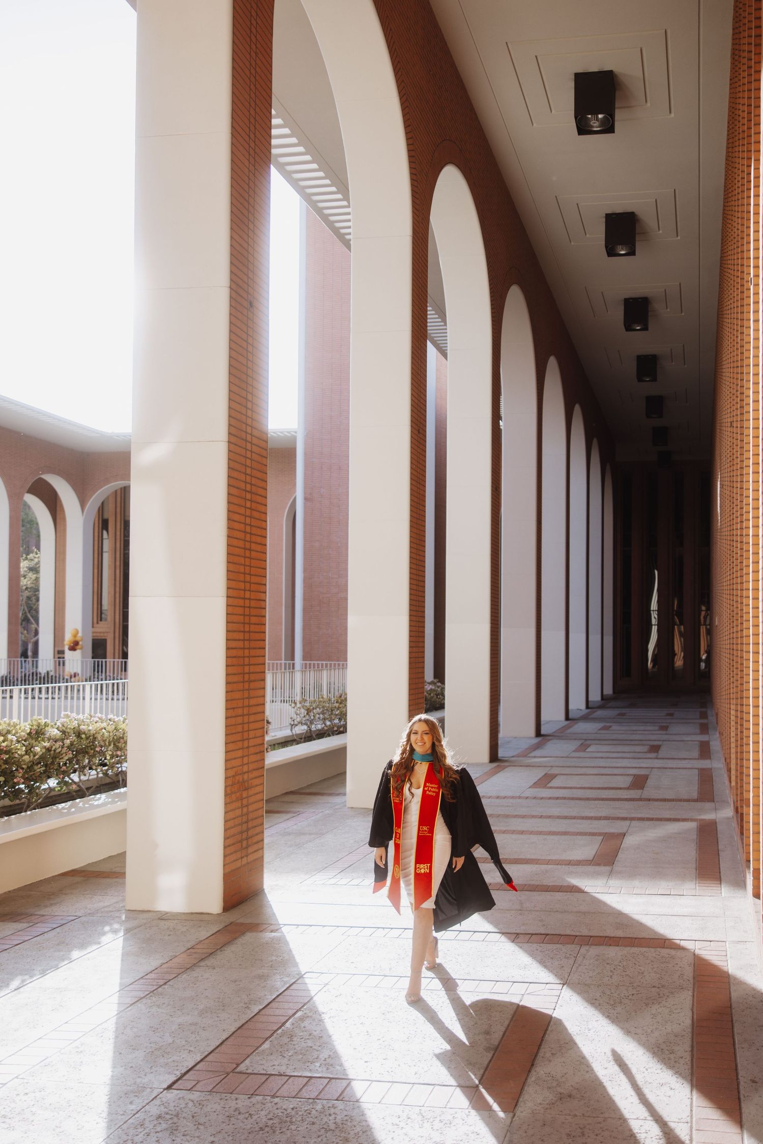 Graduate walks through classical archway corridor in graduation attire.