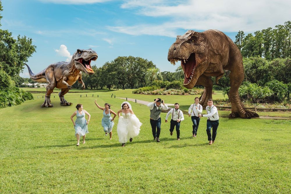 Wedding party playfully runs away from edited dinosaur photo effects on a lush green hillside.