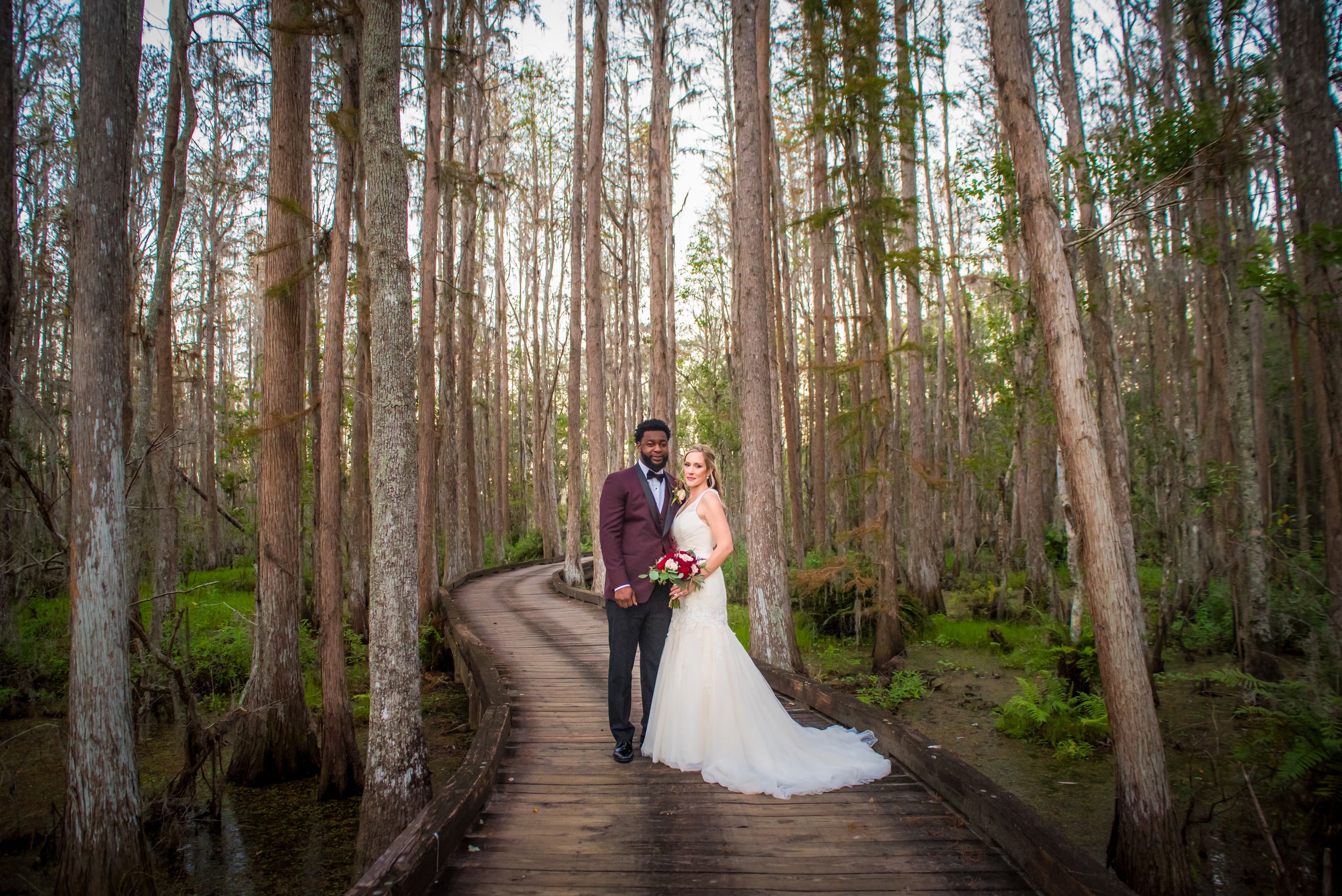 Bride and groom pose on a winding wooden boardwalk surrounded by tall trees in a lush forest setting. Tampa Wedding Photography, Tampa Photo