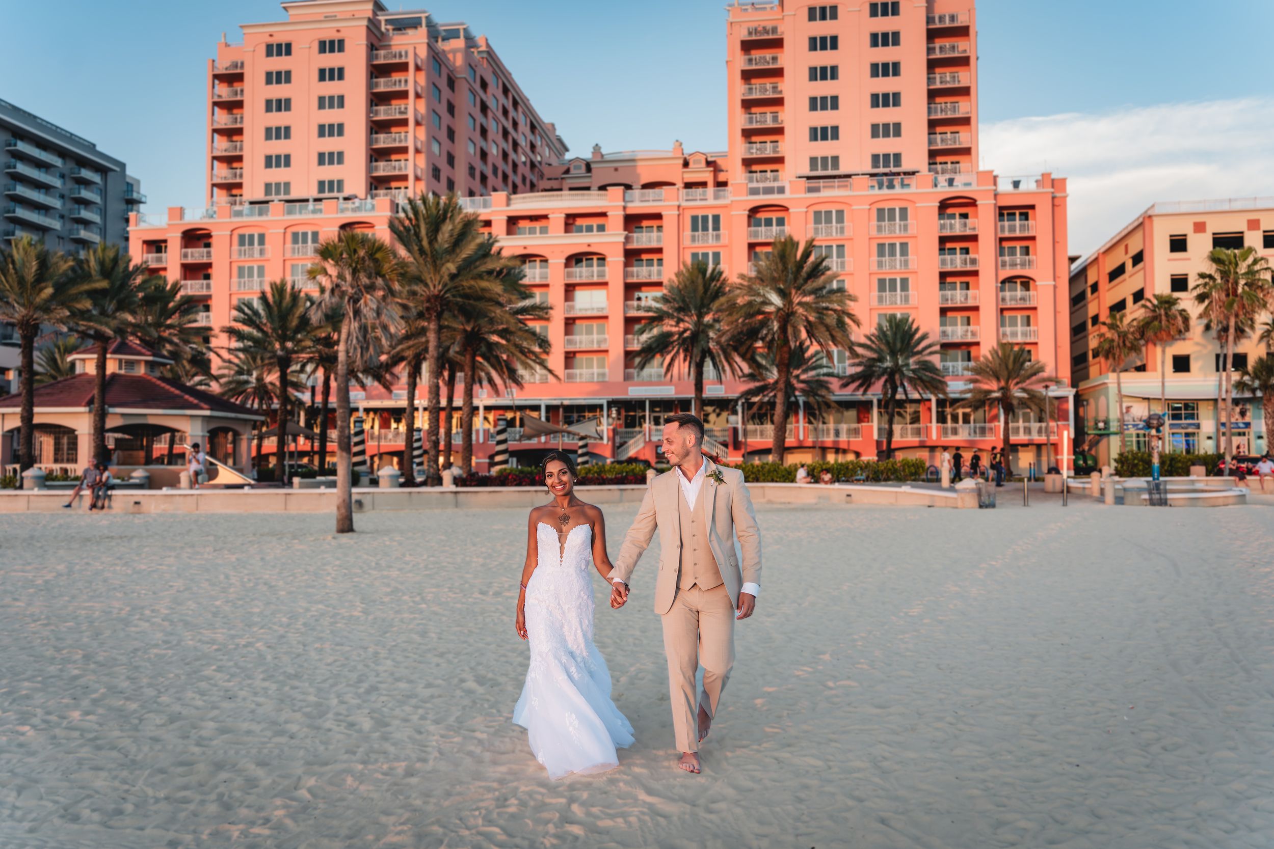 Newlywed couple holding hands on white sand beach with pink resort hotel and palm trees at sunset. Tampa Wedding Photography, Photographer