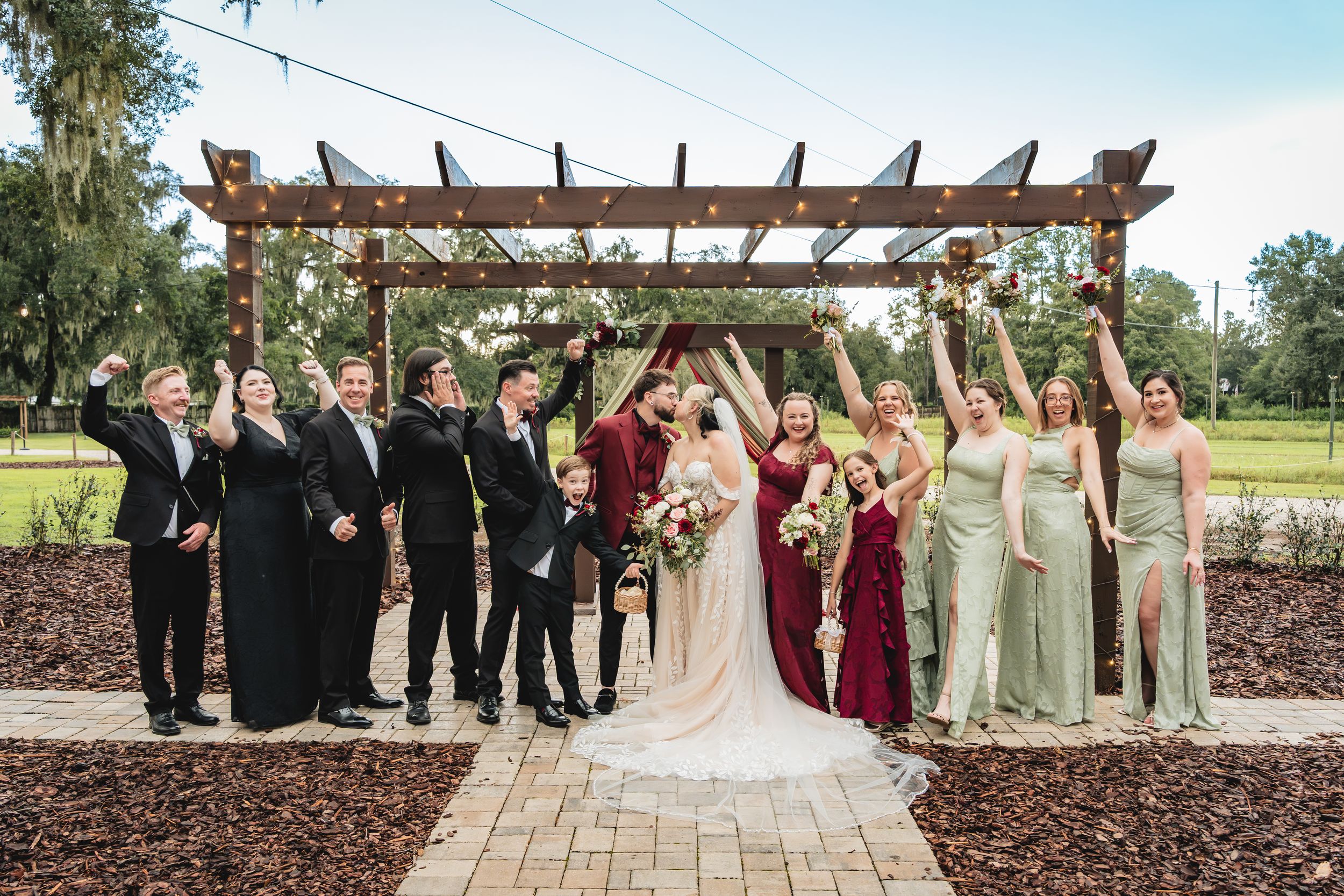Wedding party celebrating under a rustic pergola, bridesmaids in sage green and groomsmen in black, bride and groom kissing.