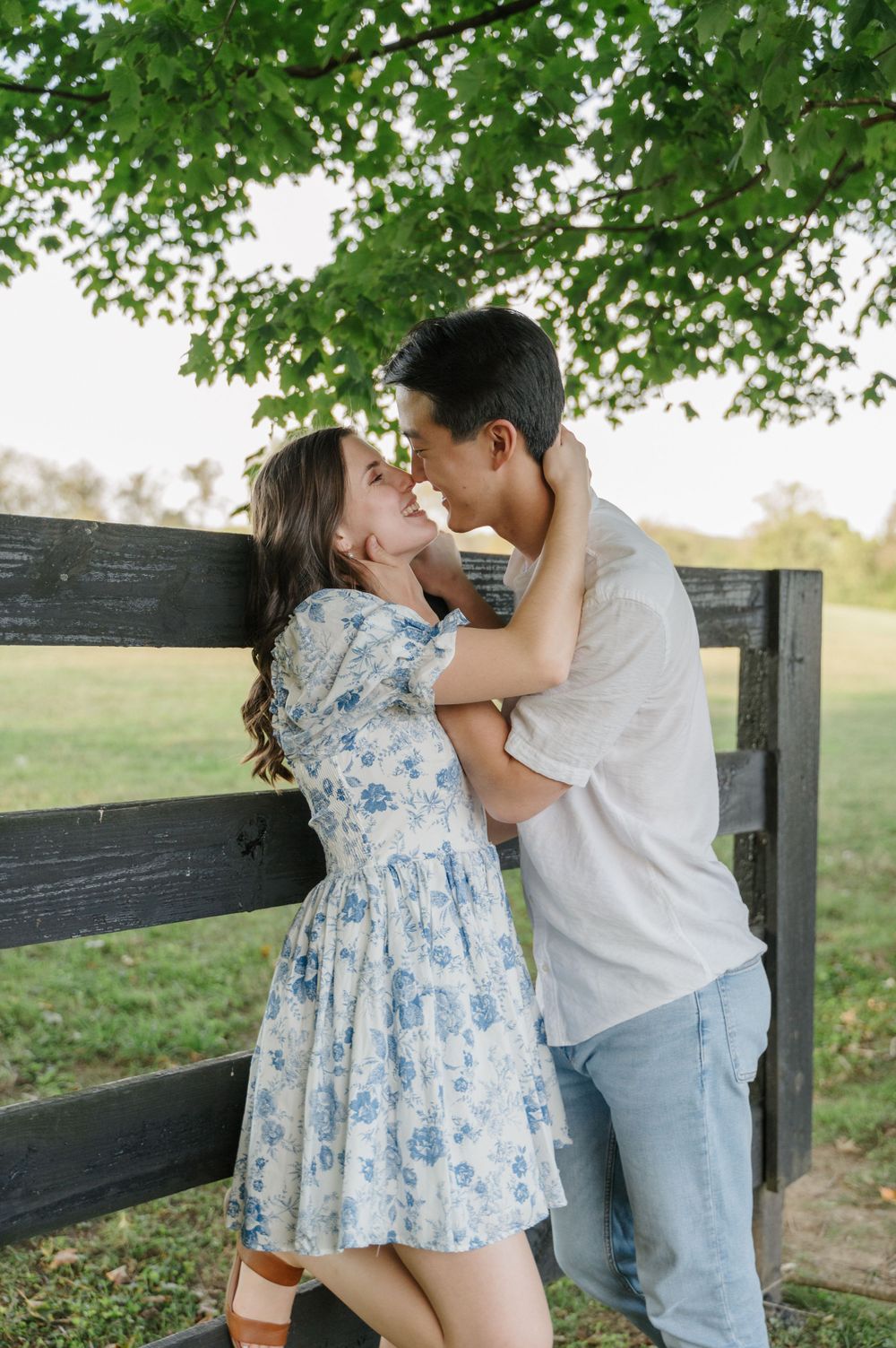 Man leans into kiss fiancé against a fence under a tree