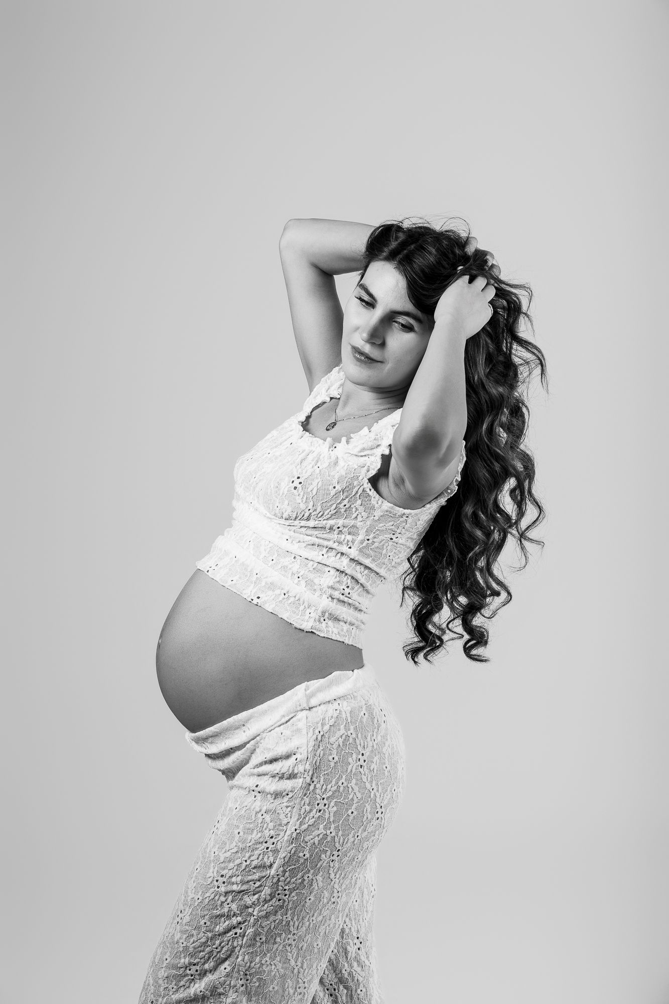 Black and white portrait of a pregnant woman with long curly hair posing gracefully in a white lace maternity outfit.