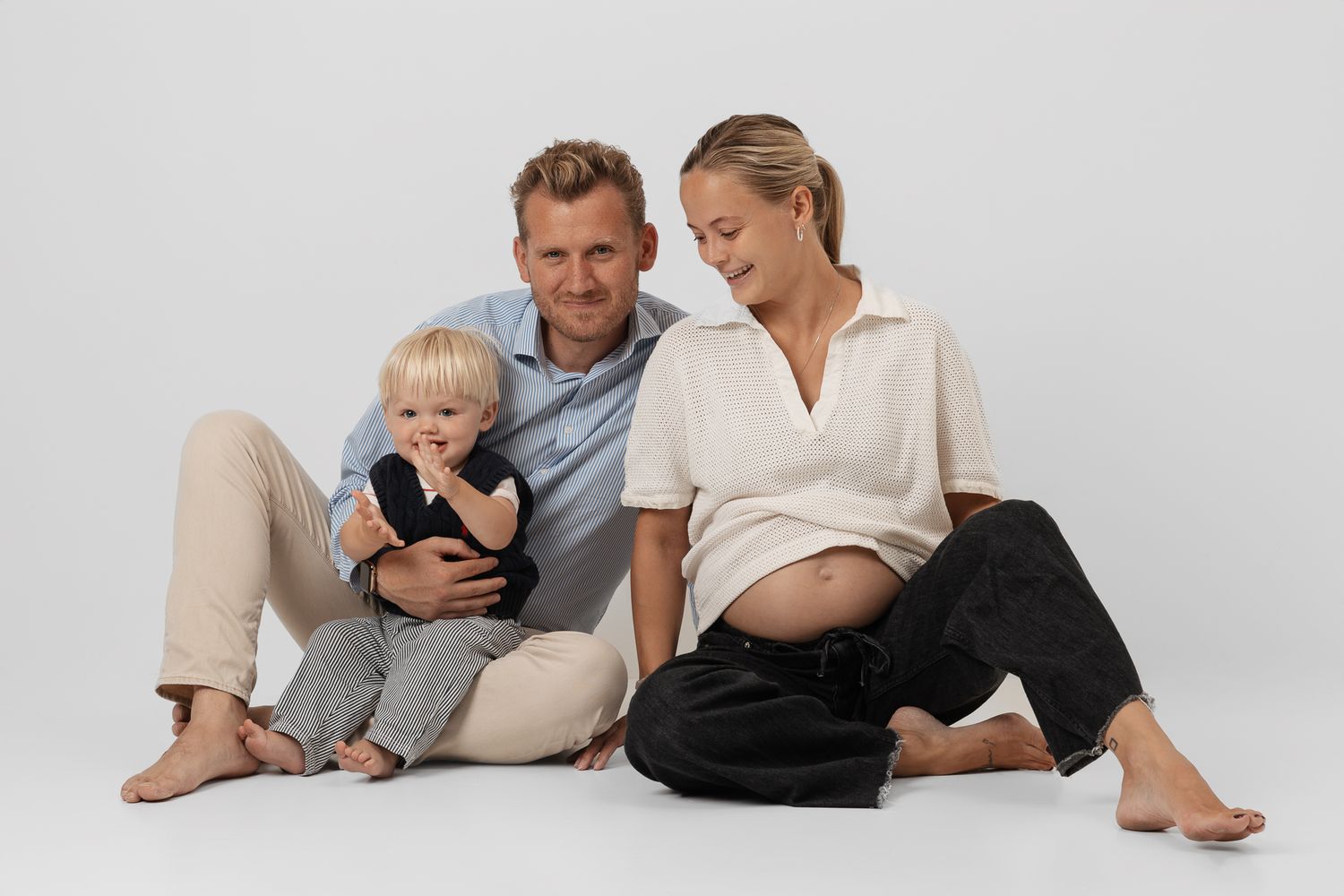 A family lounges comfortably on white bedding while using mobile devices together.