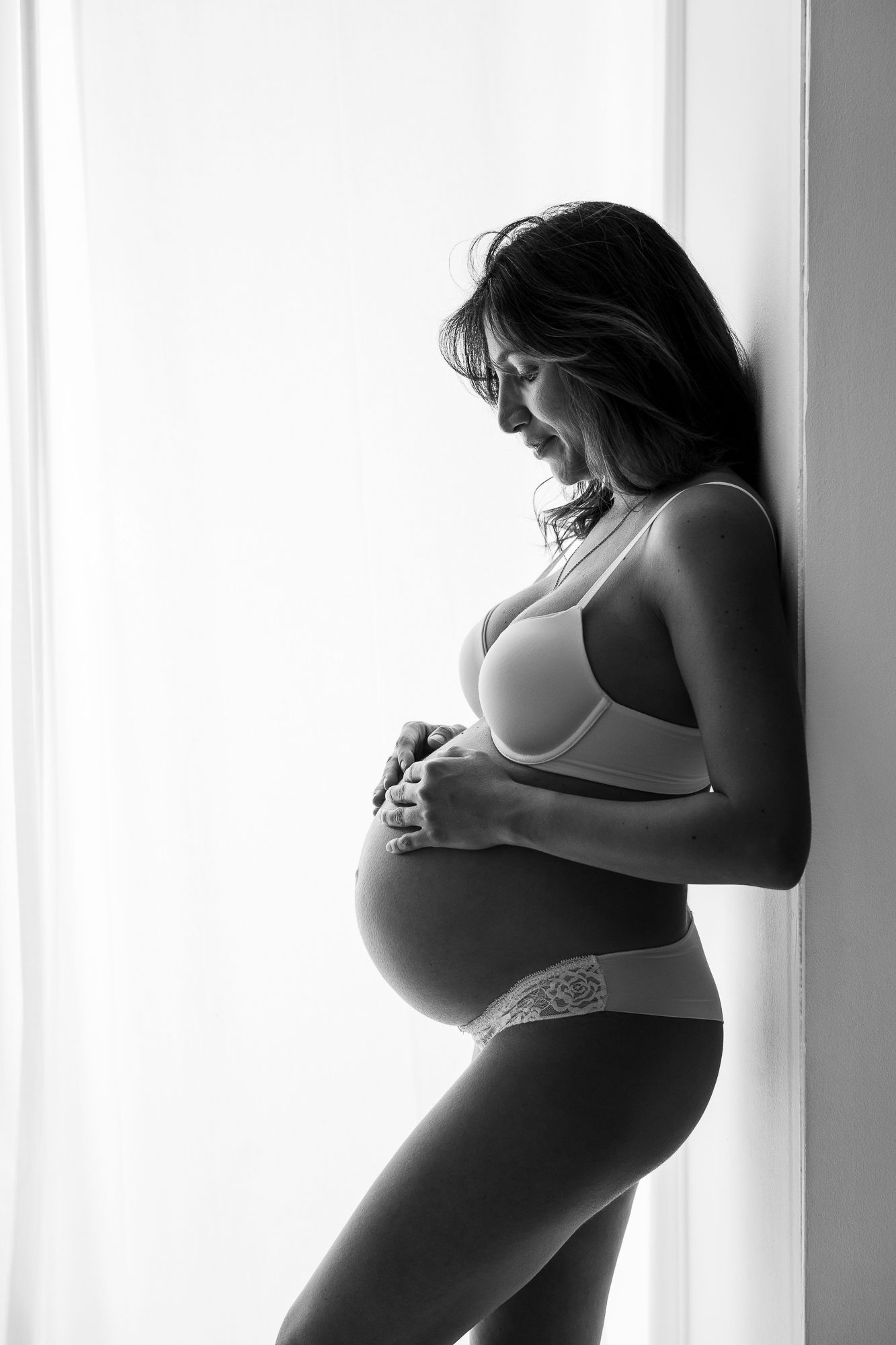 Black and white photo of a smiling pregnant woman in lingerie cradling her baby bump by a bright window.