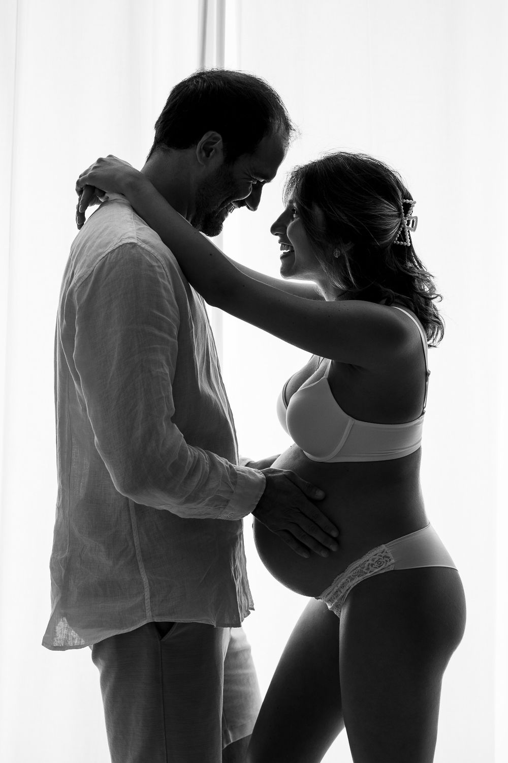 Black and white photo of a couple sharing a tender moment, the woman visibly pregnant, smiling at each other near a window.