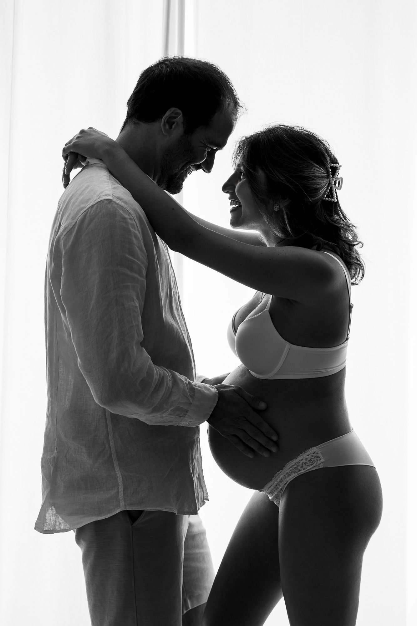 Black and white photo of a couple sharing a tender moment, the woman visibly pregnant, smiling at each other near a window.