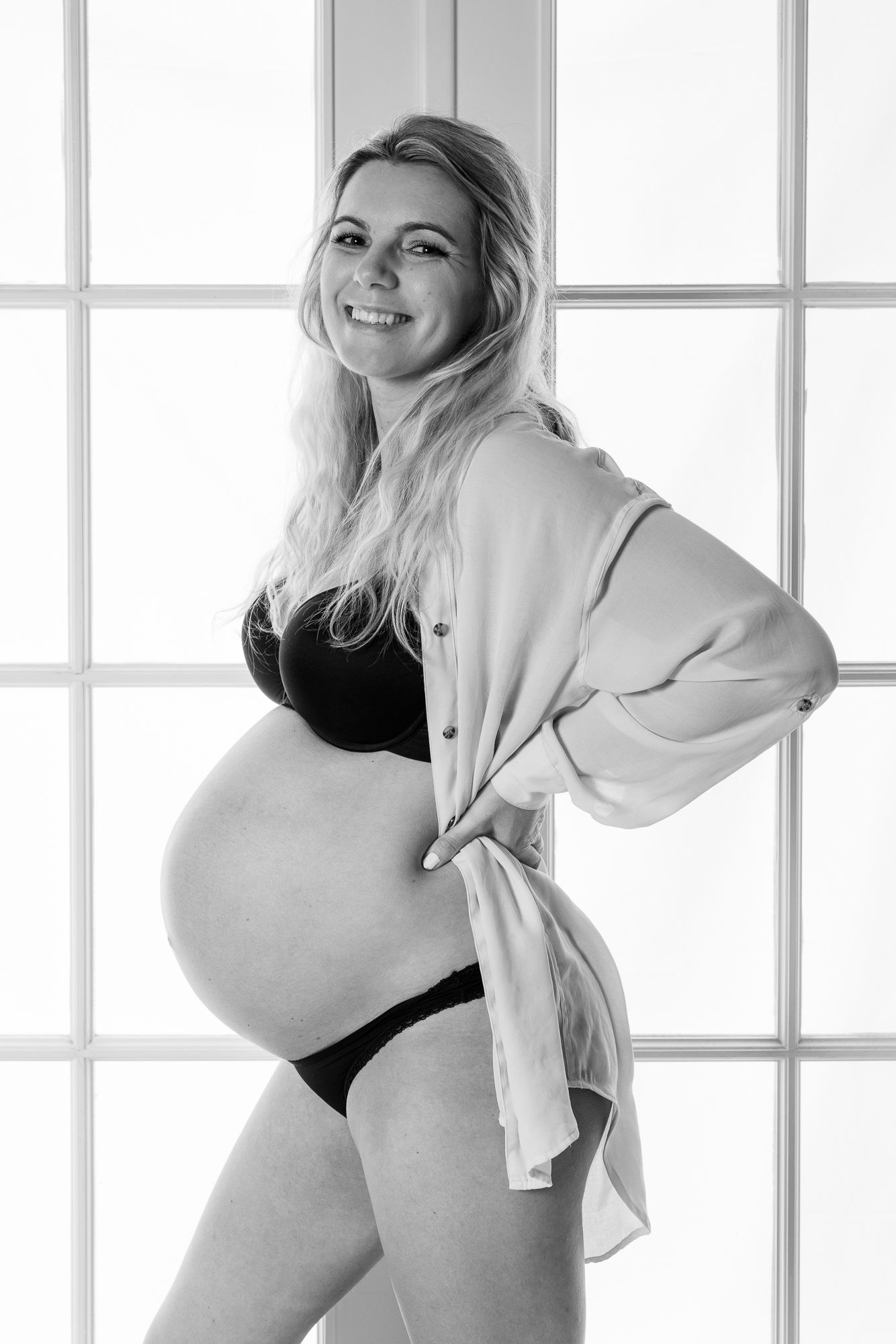A black and white maternity portrait shows a woman by a window in undergarments and an open button-down shirt.