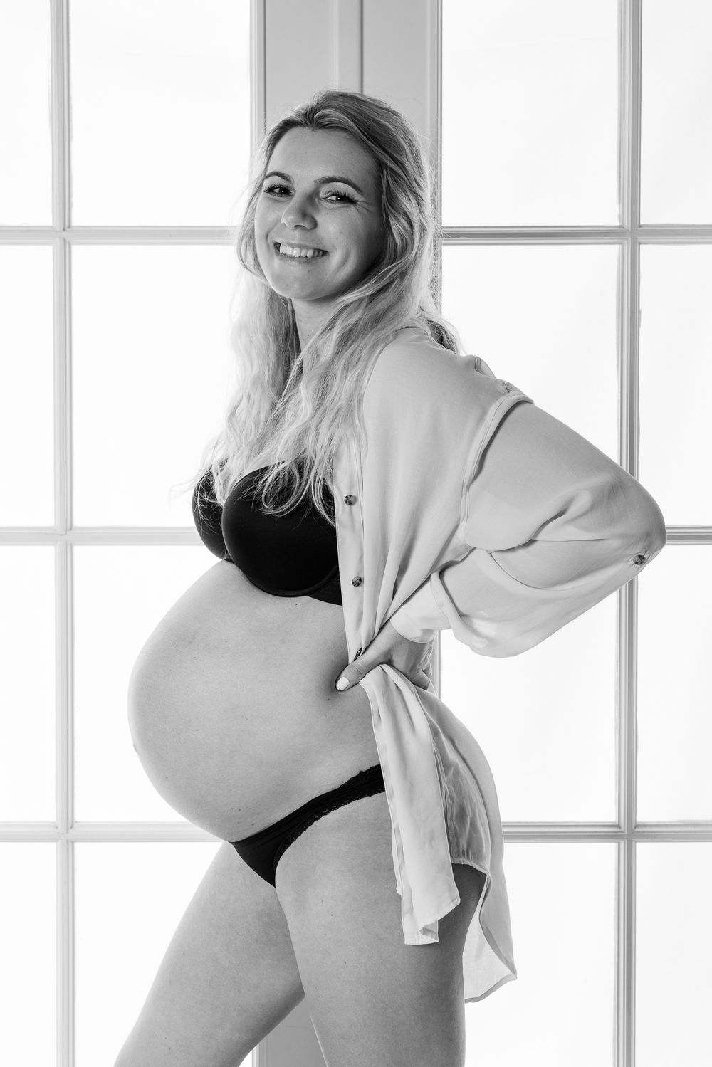 A black and white maternity portrait shows a woman by a window in undergarments and an open button-down shirt.