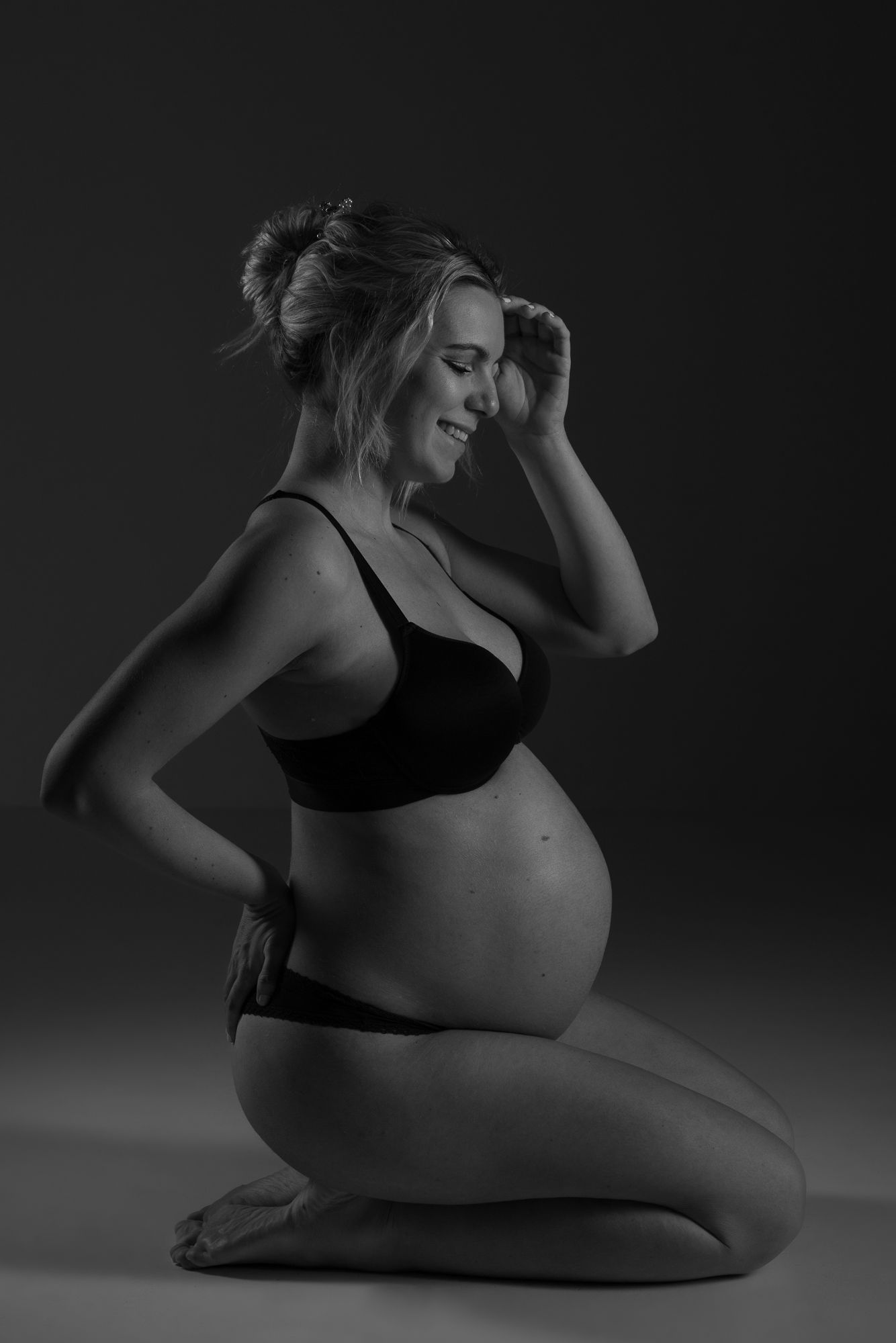 Artistic black and white maternity portrait showing a pregnant woman in intimate apparel kneeling in profile.