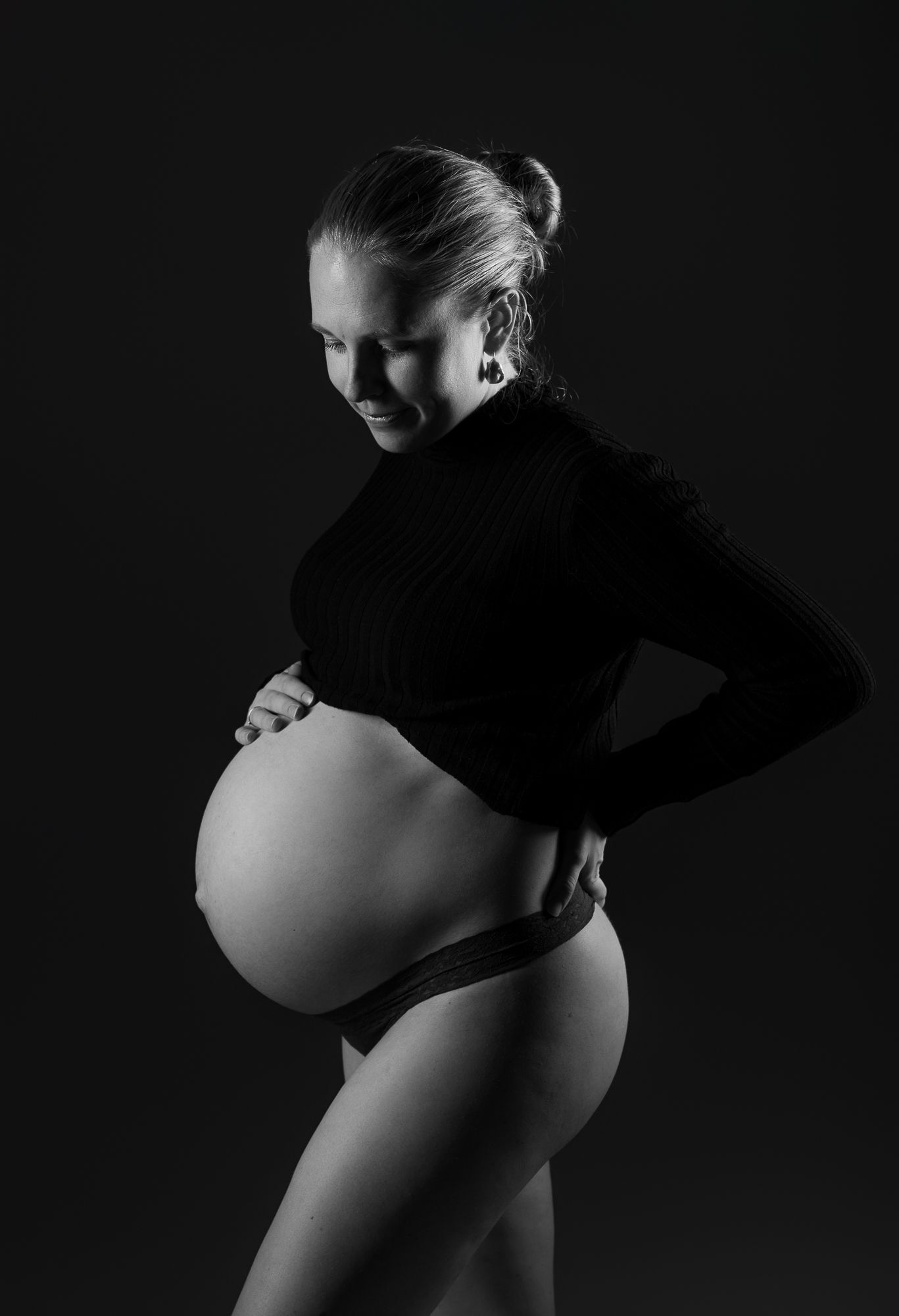 Artistic black and white maternity photo showing silhouette of pregnant woman in profile against dark background.