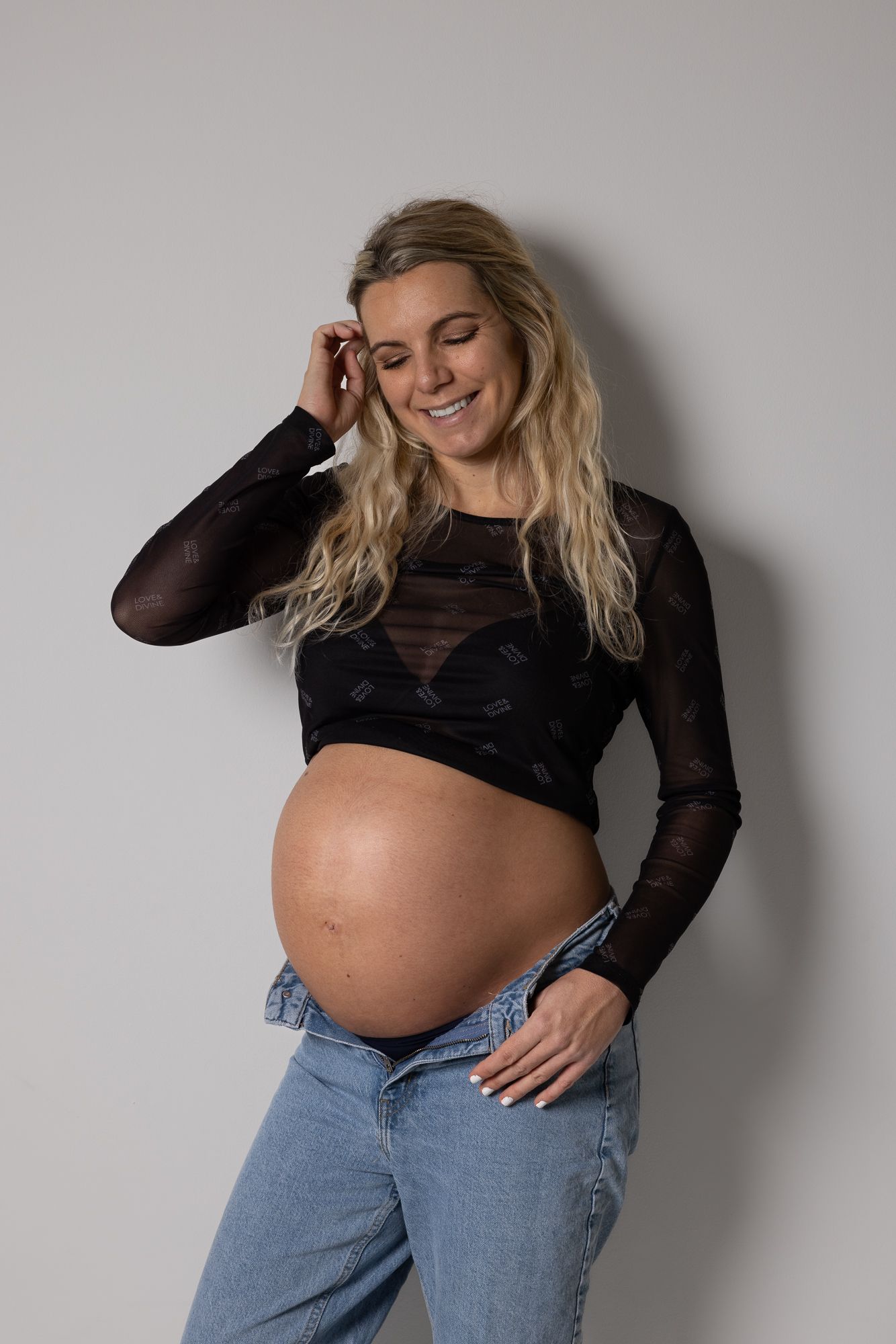 Expectant mother in black sheer top and jeans poses against gray wall with hand on pregnant belly.