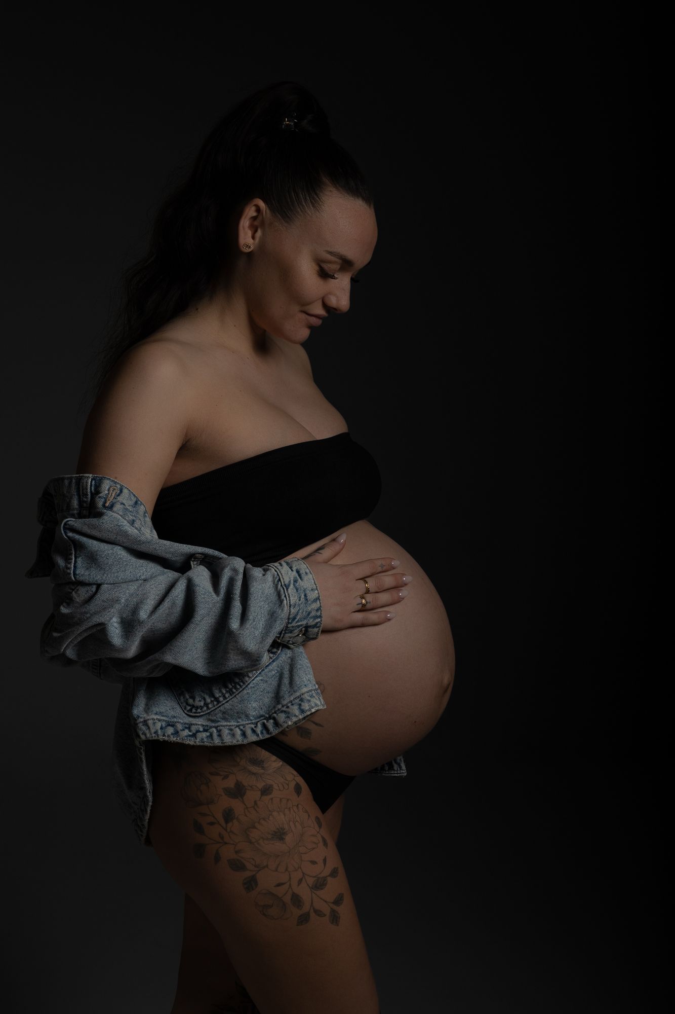 Artistic maternity photo with subject in black top and denim jacket against dark background showing pregnant belly.