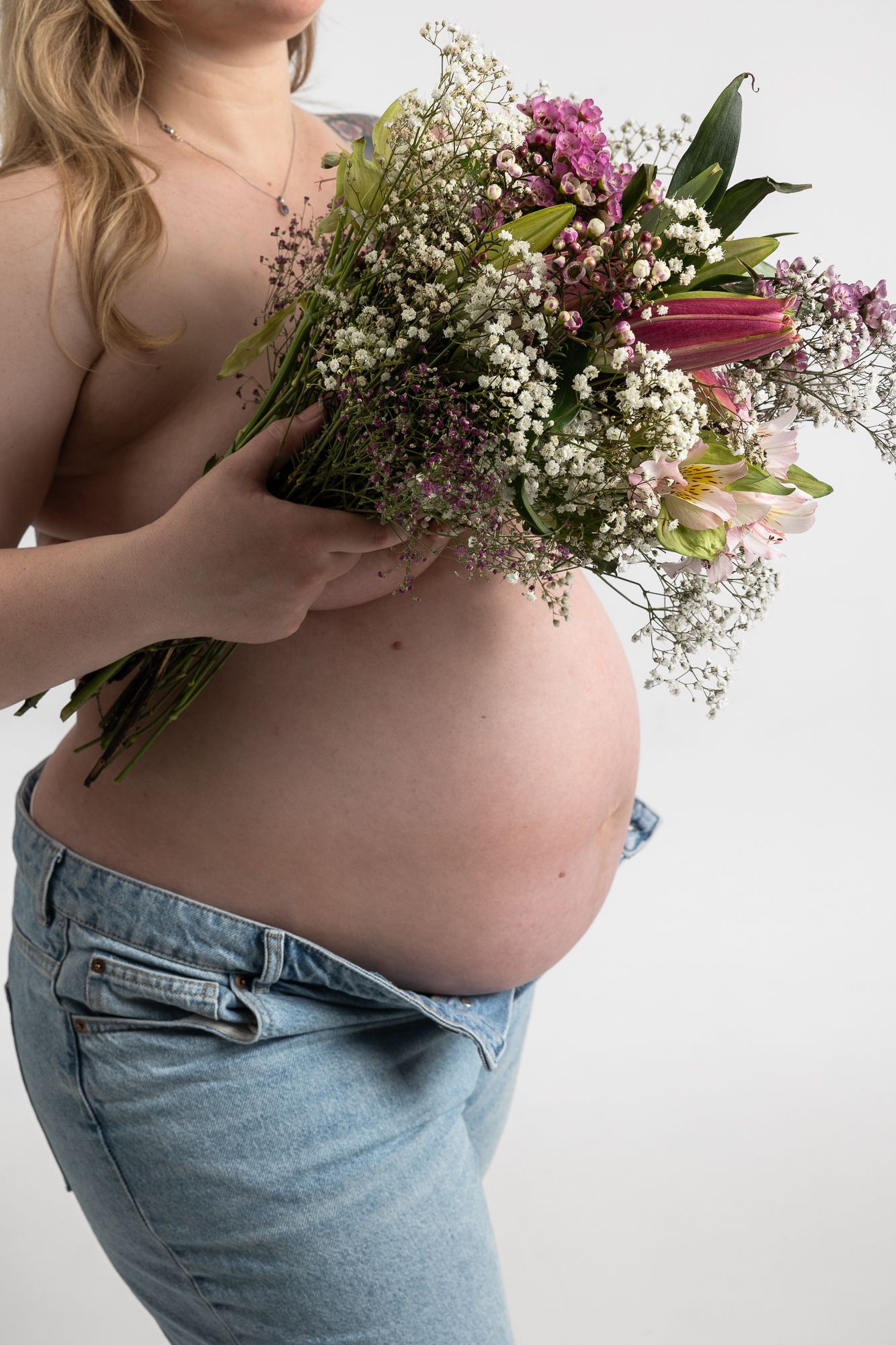 A pregnant belly shown in profile with hands holding a delicate bouquet of baby's breath and pink flowers against white background.