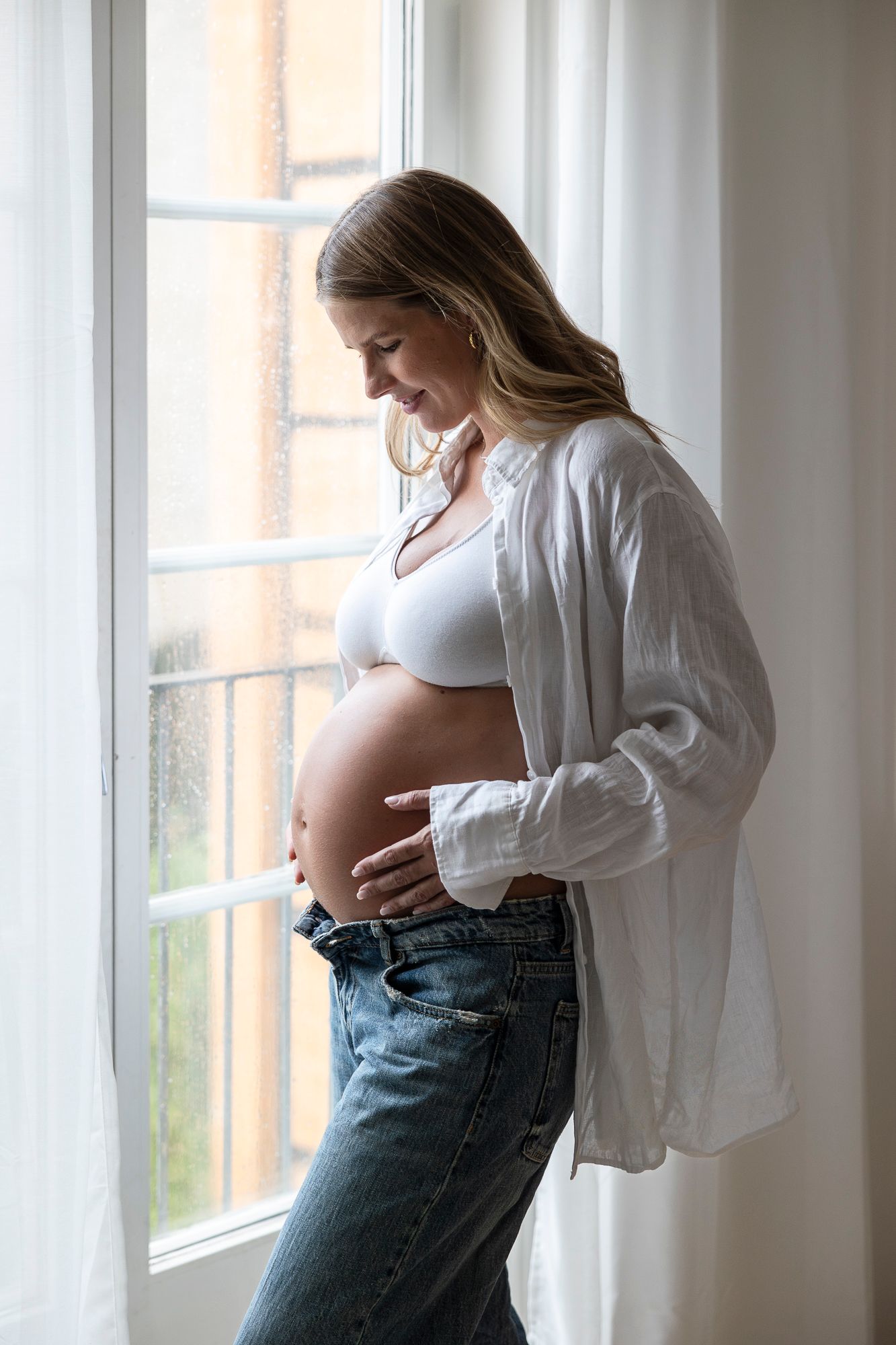 Expectant mother in white shirt and jeans stands by window, lovingly cradling her pregnant belly in natural light.