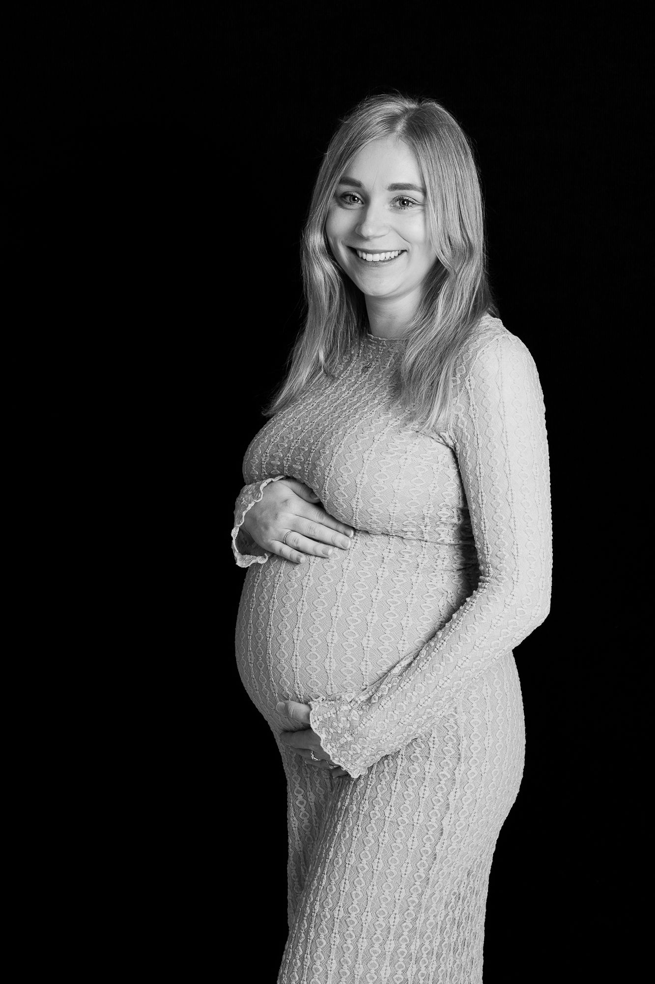Black and white maternity portrait of an expectant mother wearing a knit dress and smiling against dark background.